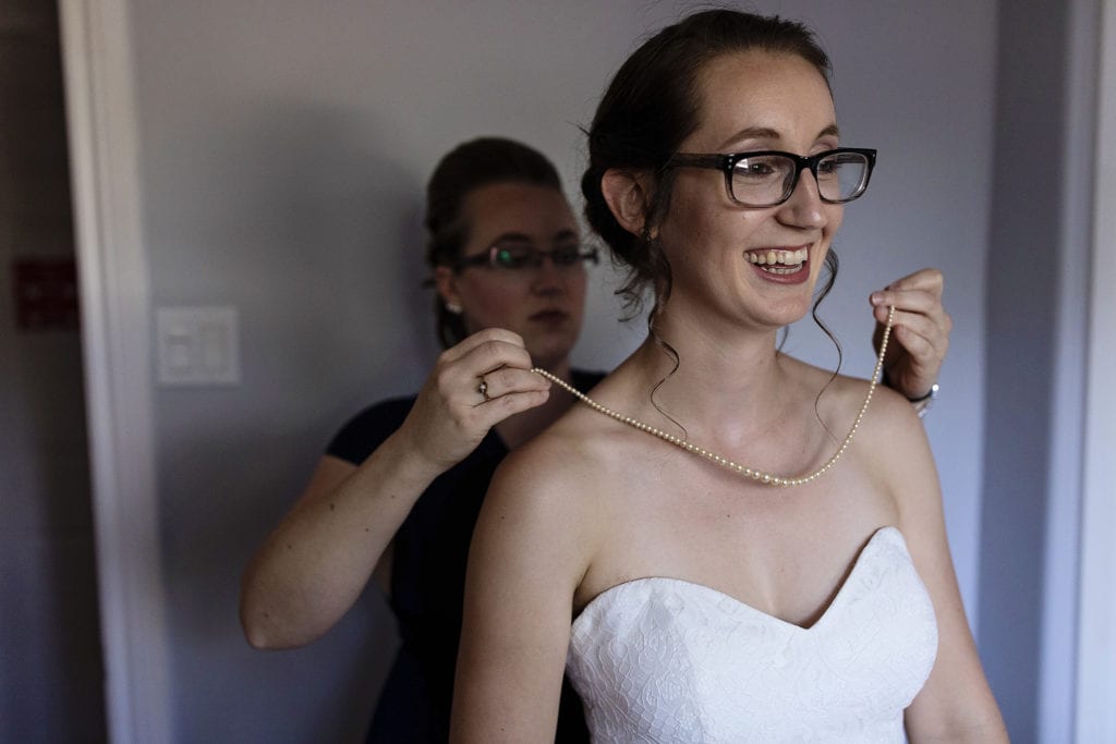 bride smiles as sister puts pearl necklace on her