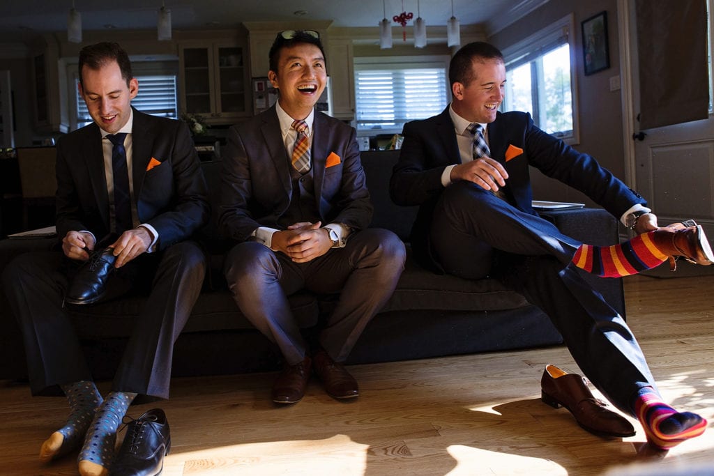 groom laughs while groomsmen put on socks and shoes on couch