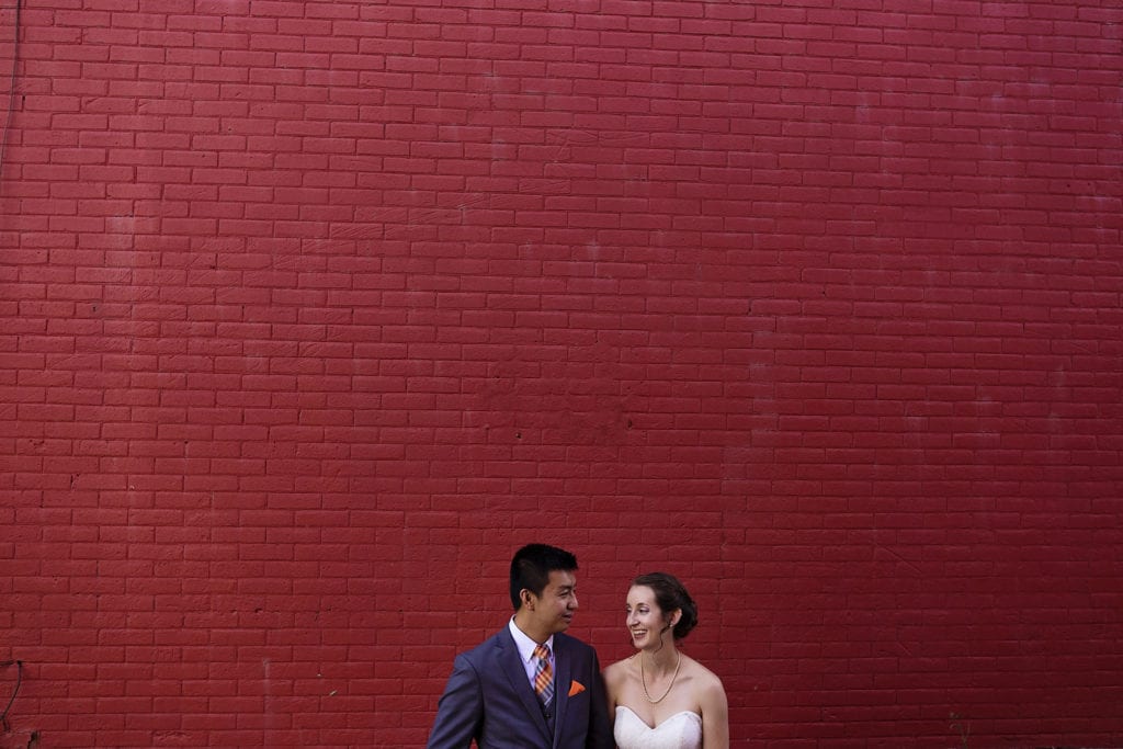 bride and groom look past each other in front of red wall