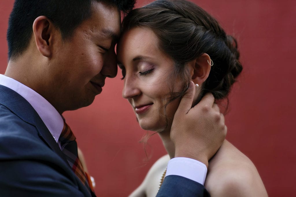 groom holds bride's face close and rests forehead against her while standing in front of red wall