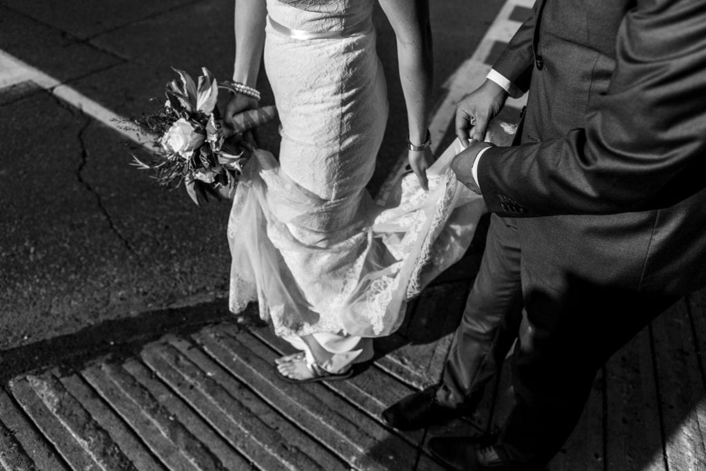 bride and groom remove dirt from hem of bride's dress on sidewalk