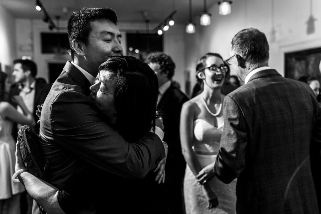 groom hugs mother and closes his eyes as bride dances with father Orange Art Gallery wedding reception