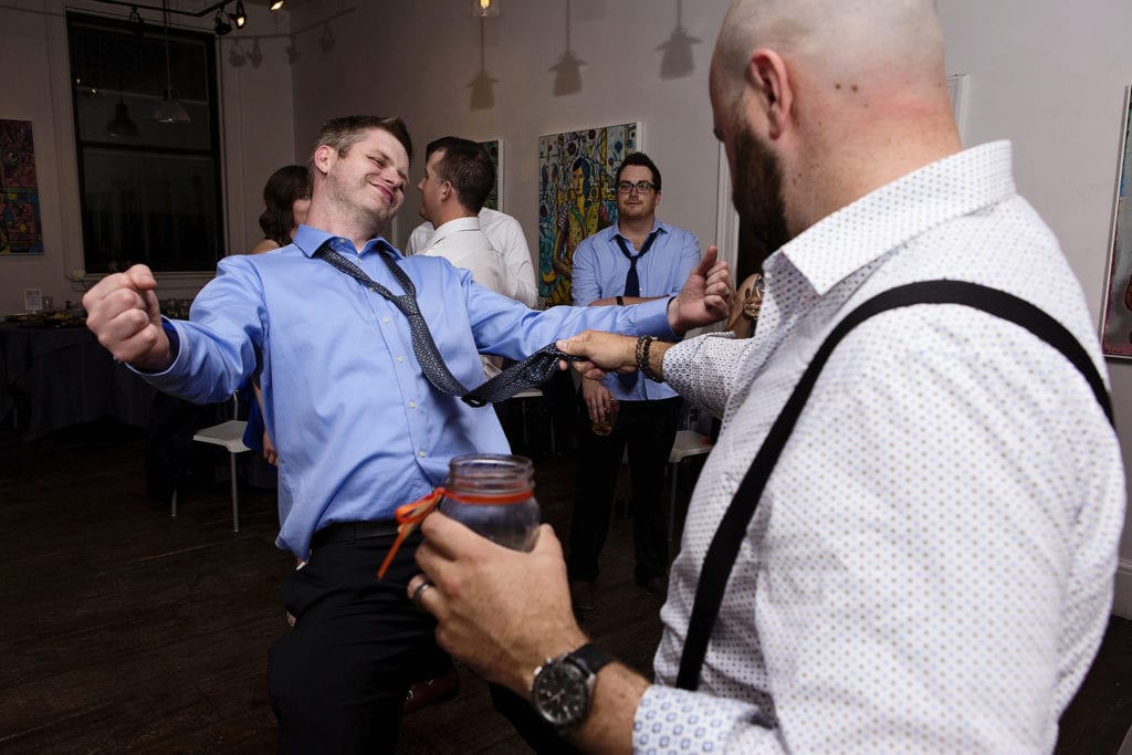 man dances with arms up as other man holds his tie