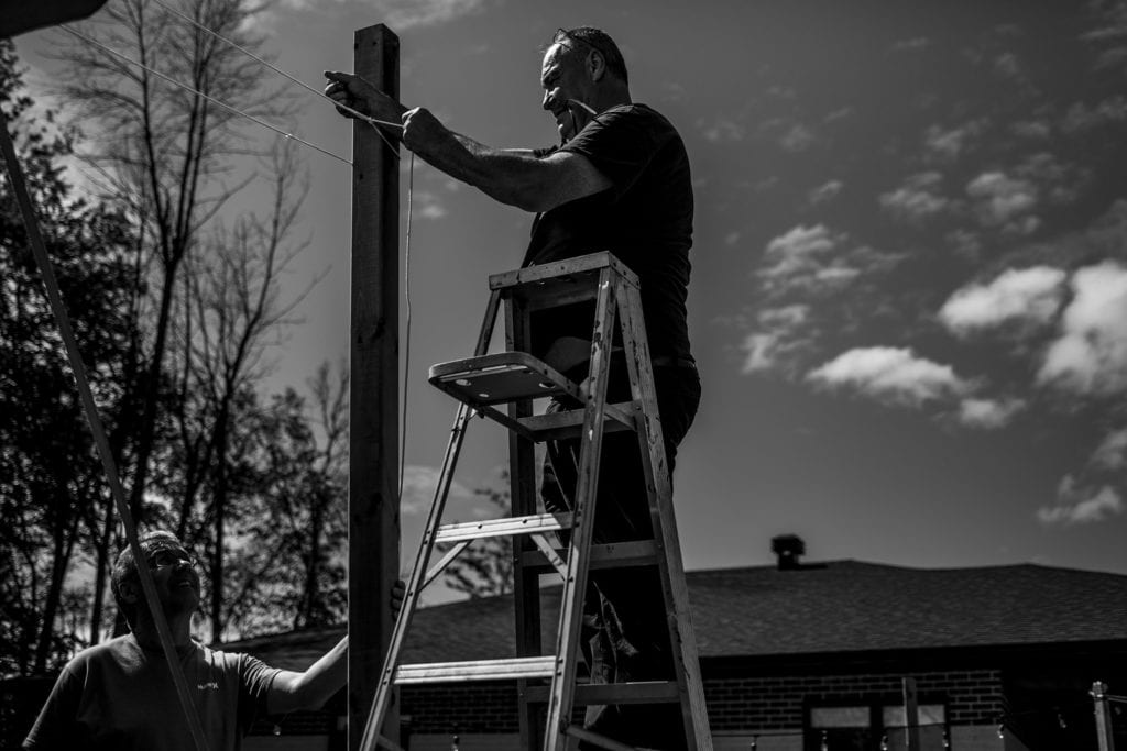 man stands on ladder and holds tool in mouth while fastening rope to pole