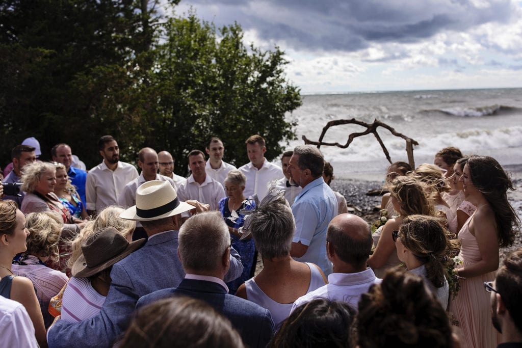 guests cluster together at shorefront for windy Point Petre wedding ceremony while waves roll in background