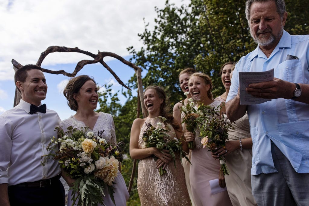 bridesmaids laugh at smiling bride and groom while officiant conducts ceremony