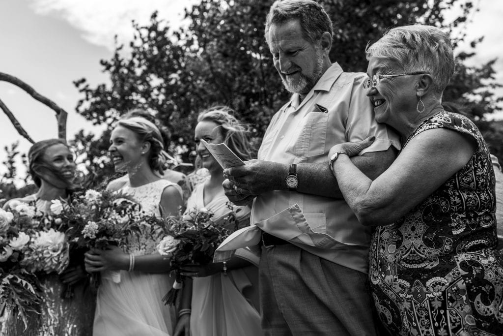 female officiant wraps ars around husband while he conducts wedding ceremony and bridesmaids watch