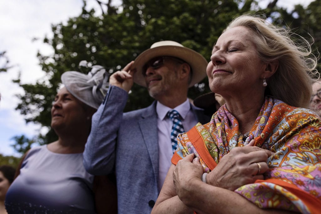 woman holds scarf around her shoulders and faces the wind with eyes closed and a smile while other wedding guests watch on