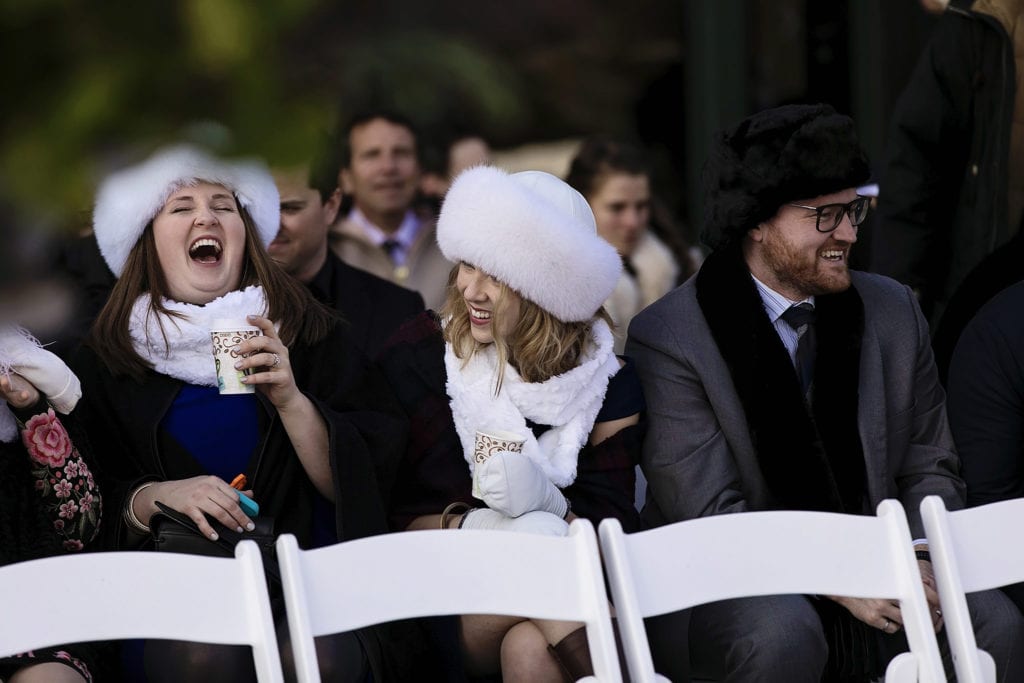 wedding guests in white fur hats laugh while seated in white wedding chairs