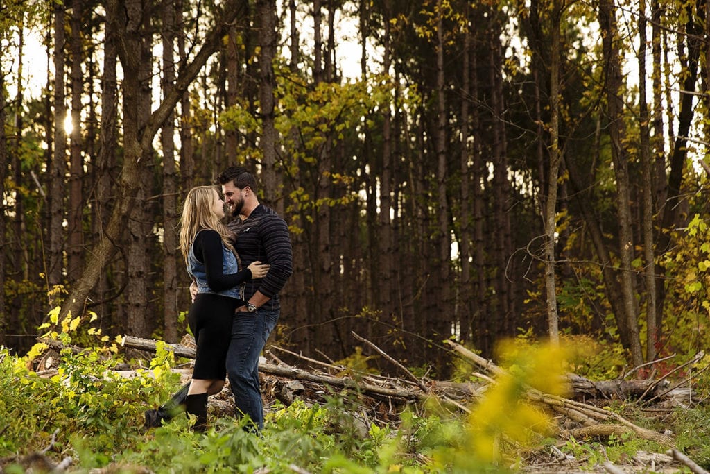 woman in blac dress and denim vest with man in striped shirt in forest during woodsy engagement shoot