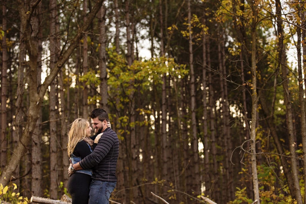 woman in black dress and denim vest with man in striped shirt embracing in forest during woodsy engagement shoot