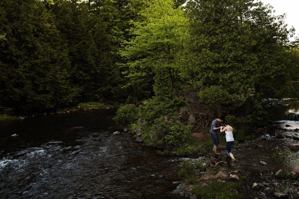 man helping woman wade across shallow stream in Gatineau Park engagement session