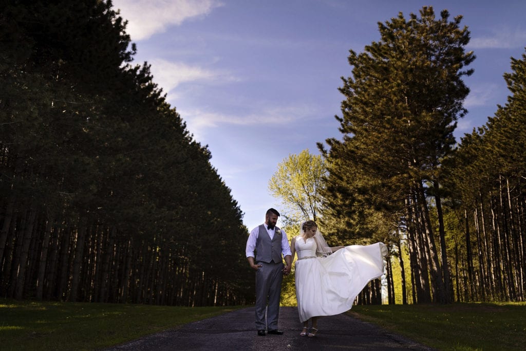 bride and groom stand in front of treeline holding hands on gravel driveway while she flares her dress out to the side