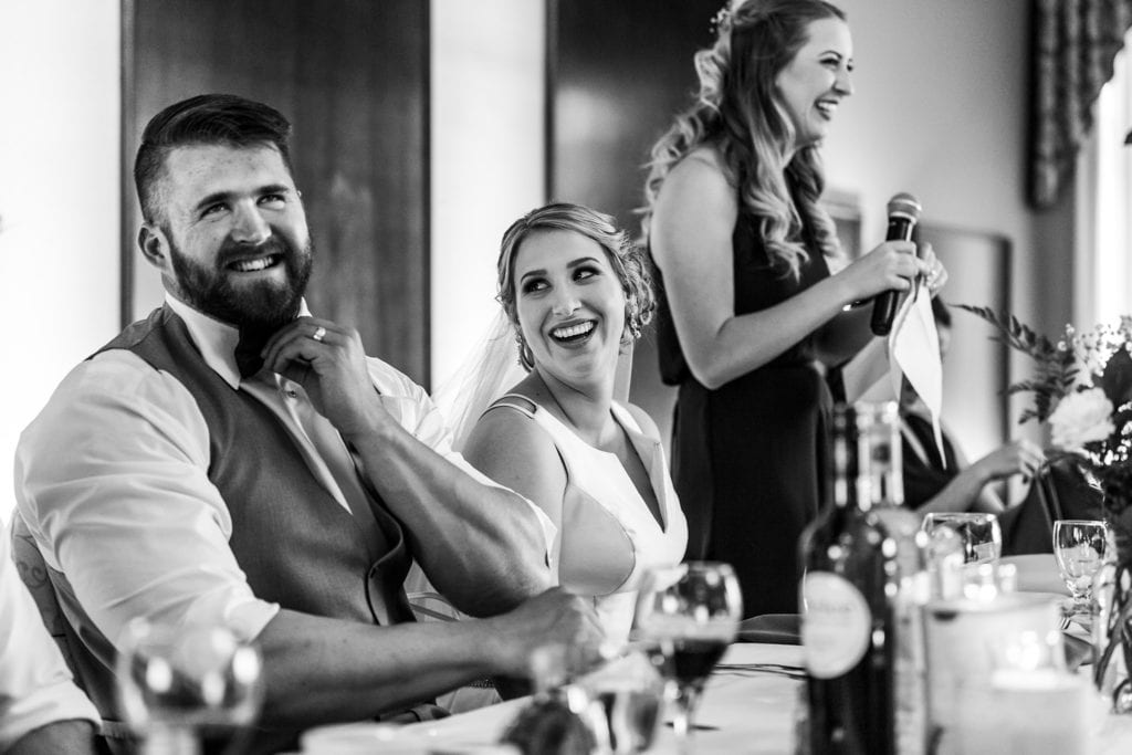 groom tugs collar and looks uncomfortable while laughing during bridesmaid's speech