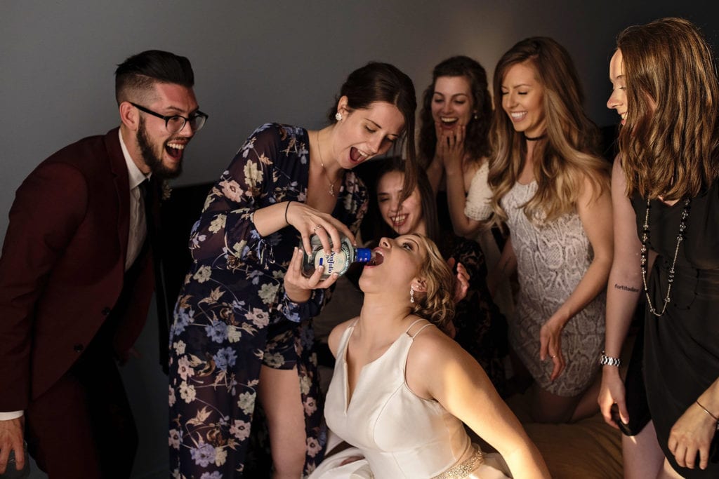 friends laugh while bride has tequila poured into her mouth straight from the bottle