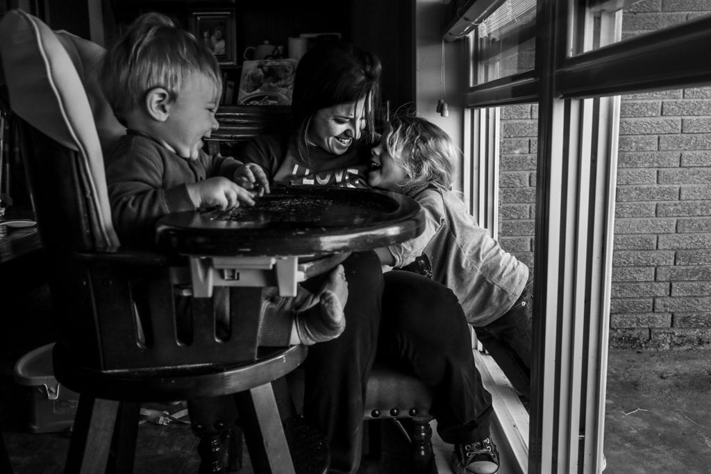 daughter leans in against mom through open window while baby laughs