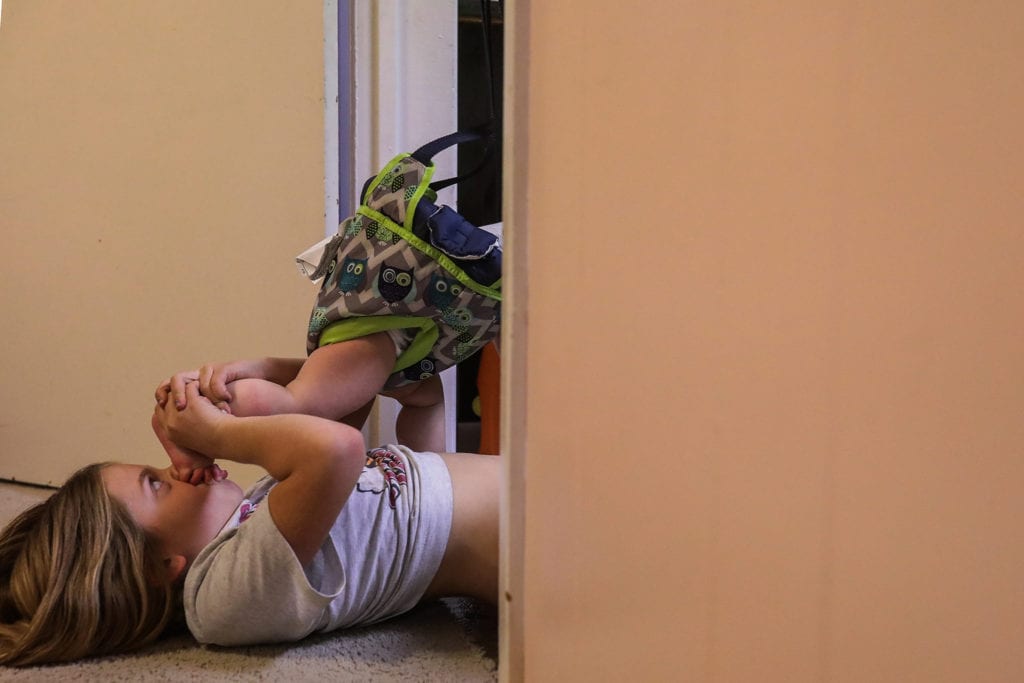 girl lying on floor kisses brother's feet while he sits in jolly jumper