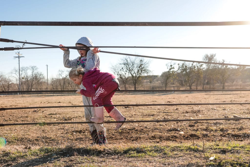 girl holds fence rail up while sister climbs under