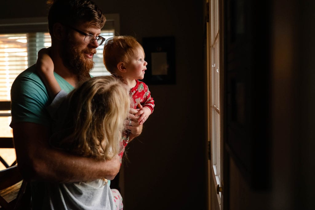Dad stands by window with baby while daughter hangs off of him