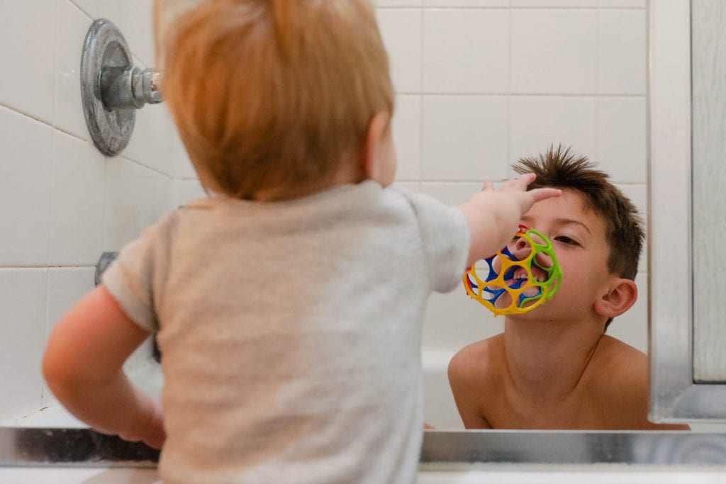 boy in bath holds toy in his mouth while baby reaches for it 