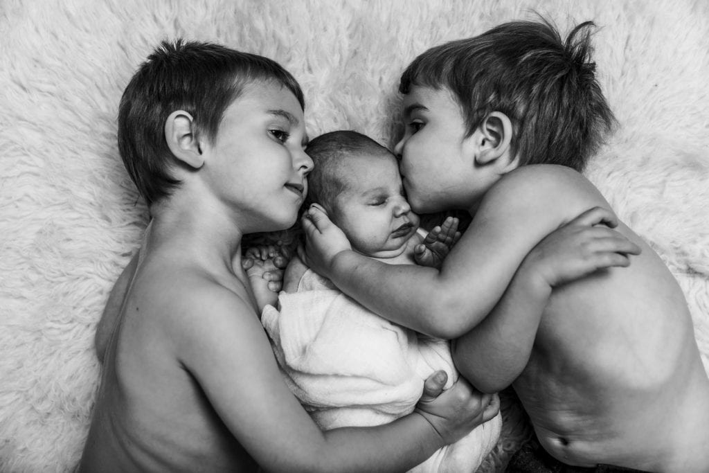 brothers lie on rug with baby sister while one kisses her during Cornwall newborn photography session