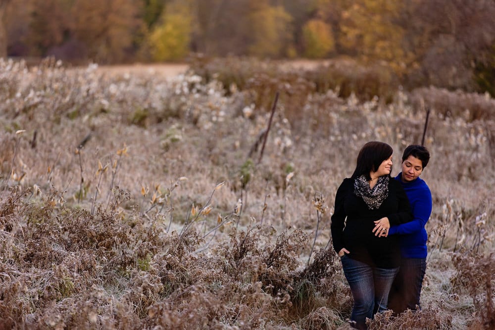 pregnant woman stands with wife in field of milkweed with hand on belly