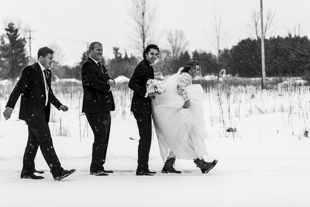 groom and groomsmen form line behind bride while she lifts her dress to show off snow boots during snowy Cornwall winter wedding