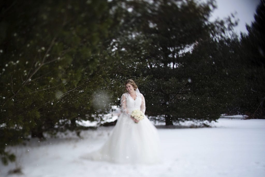 bride stands alone in snow near pine trees during snowy Cornwall winter wedding portraits