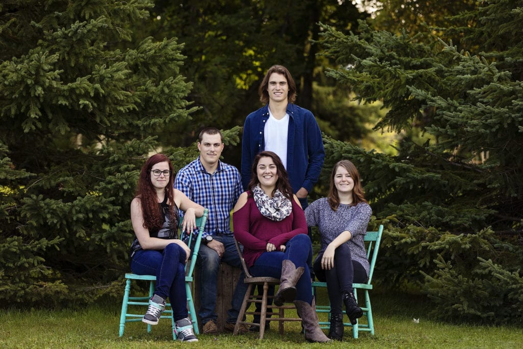 Adult children in blues and burgundies sitting on teal chairs in front of pine trees