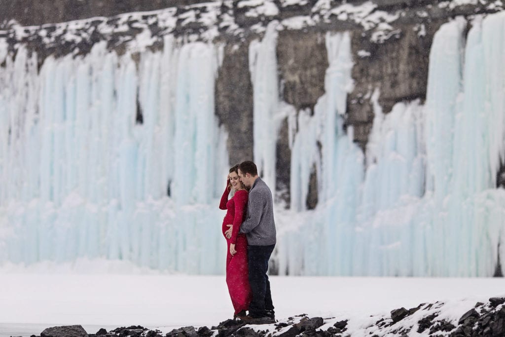 pregnant woman in red dress is held by husband with frozen waterfall in background