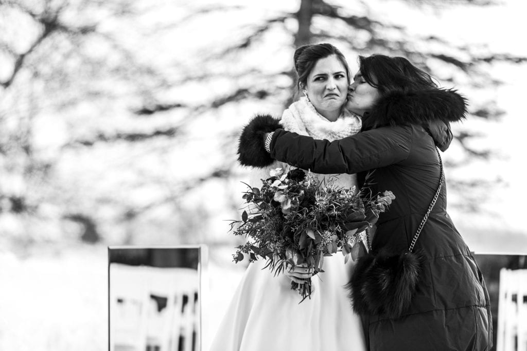 bride grimaces while holding flowers as mother kisses her cheek