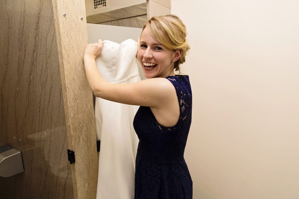 woman holds wedding dress in front of bathroom stall door