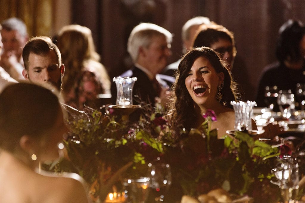 wedding guests laugh while seated at king's table with floral runner