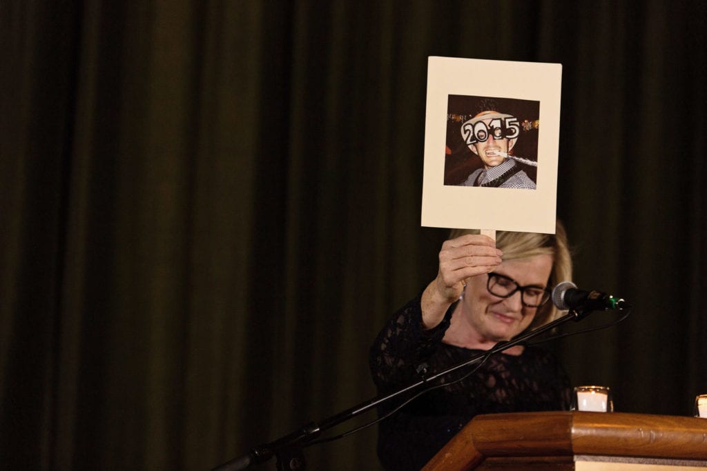 woman at podium holds up placard with photo of groom wearing funny glasses