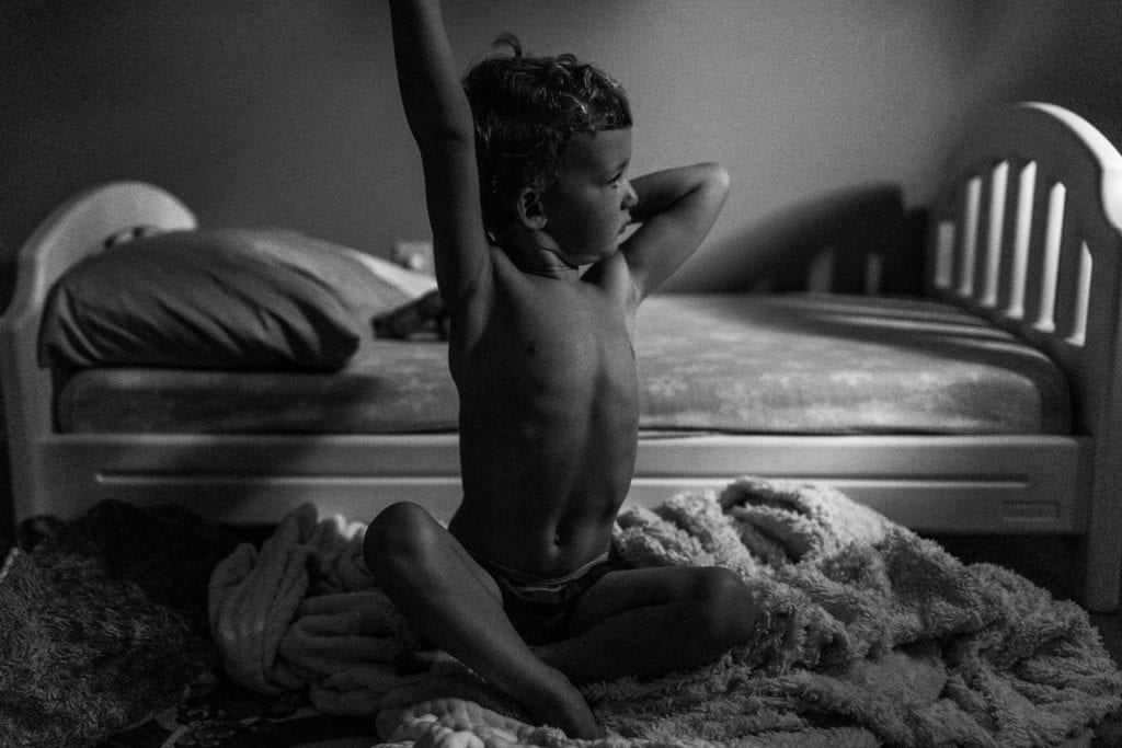 tired shirtless boy stretches while sitting on floor in front of his bed