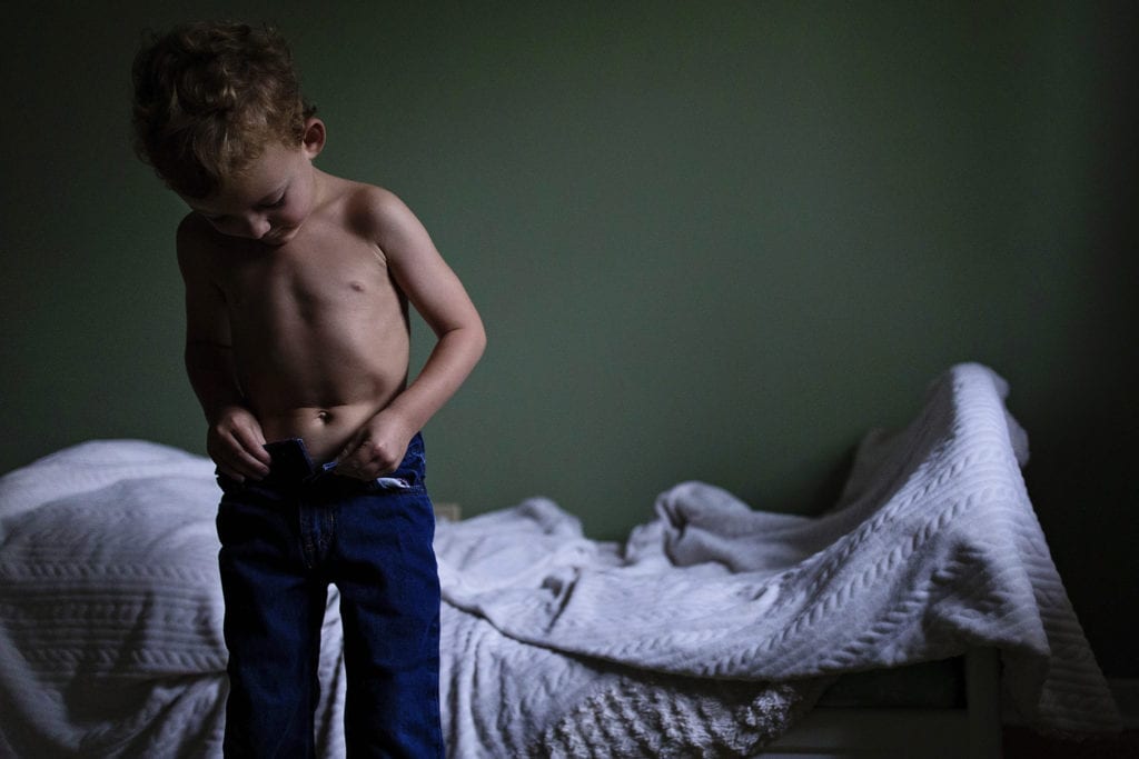 shirtless boy sucks in stomach and fastens jeans in front of his bed
