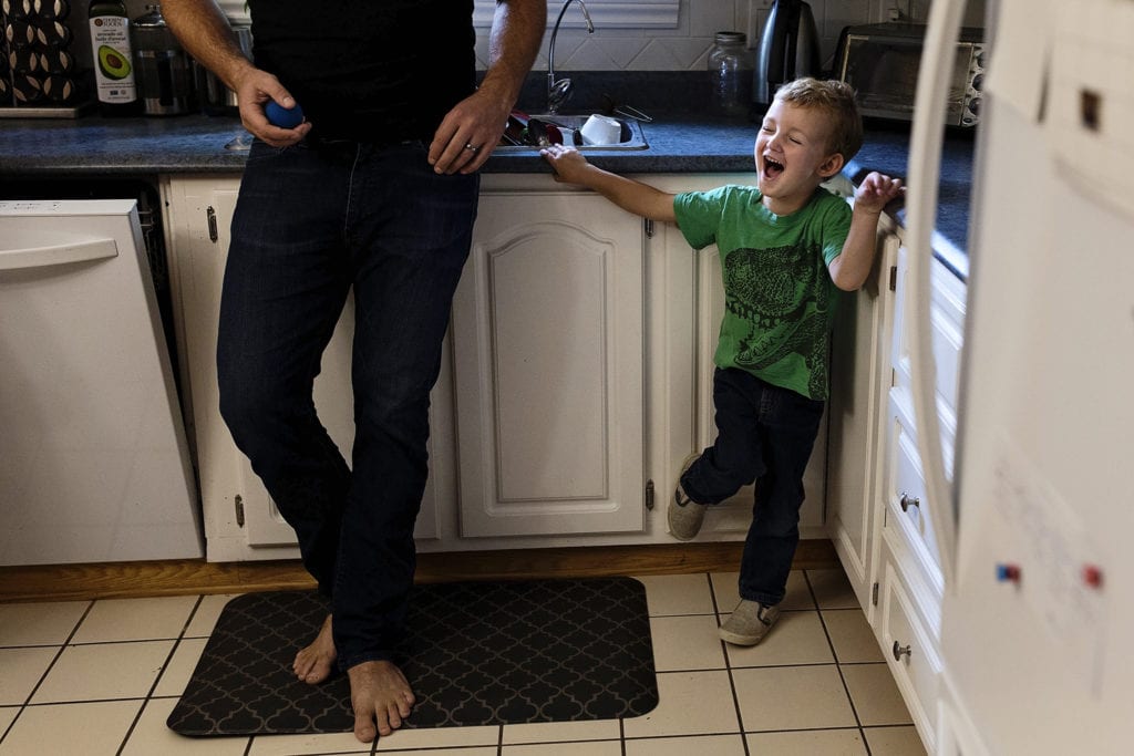 boy stands in corner of kitchen cupboards laughing while dad stands beside him