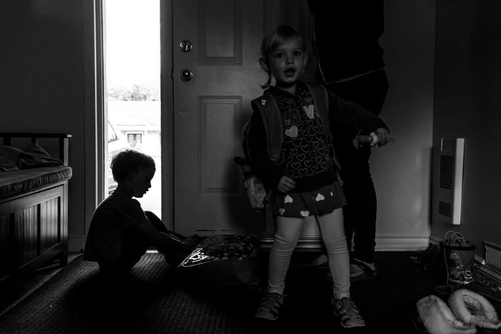 boy sits on floor to put on shoes while sister stands near door