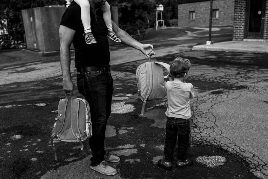 man carrying girl on shoulders and two backpacks reaches out to hand one backpack to son
