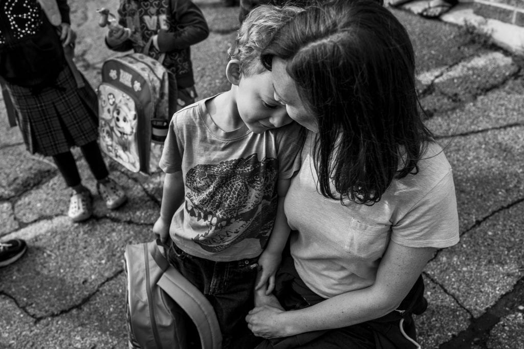 boy stands next to kneeling mom and rests head on her shoulder