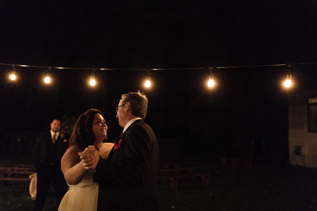 bride and uncle dance under string lights while groom watches on