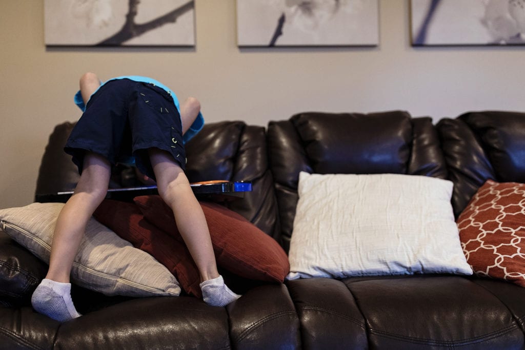 boy stands on couch with straight legs while resting head on back of couch