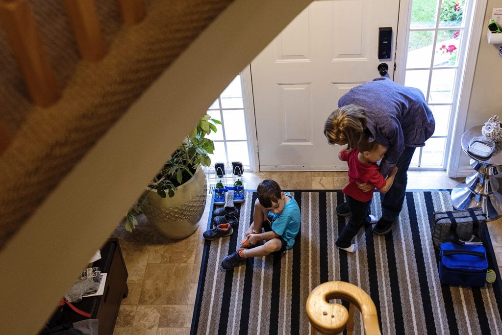 mom holds toddler while boy in blue shirt puts shoes on in front of front door