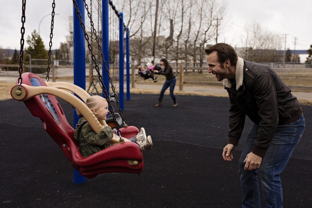 dad smiles at swinging daughter while mom and daughters play in background