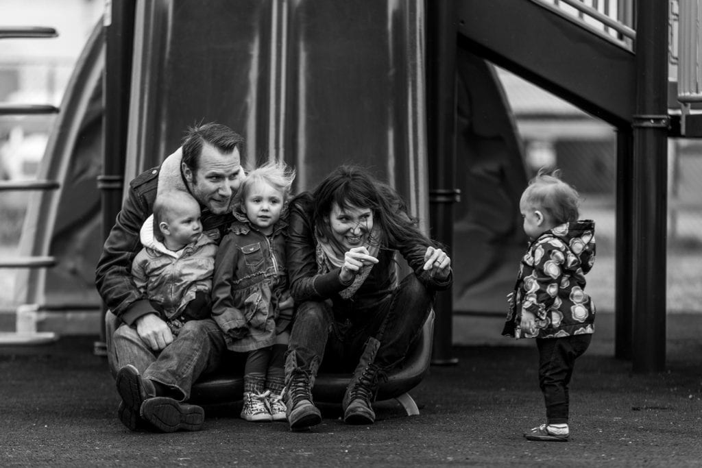 mom and dad sit with two girls on slide and make faces at third daughter