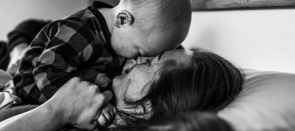 baby boy in pyjamas leans forehead against mom's forehead while lying in bed