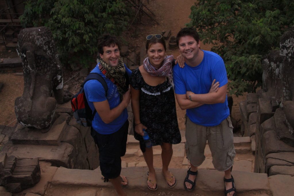two men in blue shirts stand either side of woman in black dress