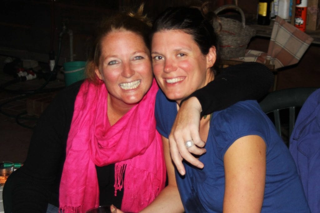 woman in black top and pink scarf sits with arm around woman in blue shirt