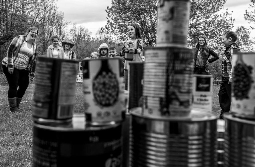 girl holds Nerf gun and aims at tin can targets while children watch