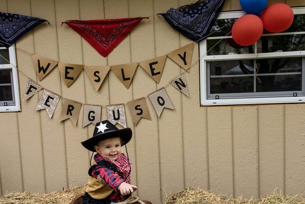 boy in cowboy outfit sits on saddle in front of bunting with his name on it